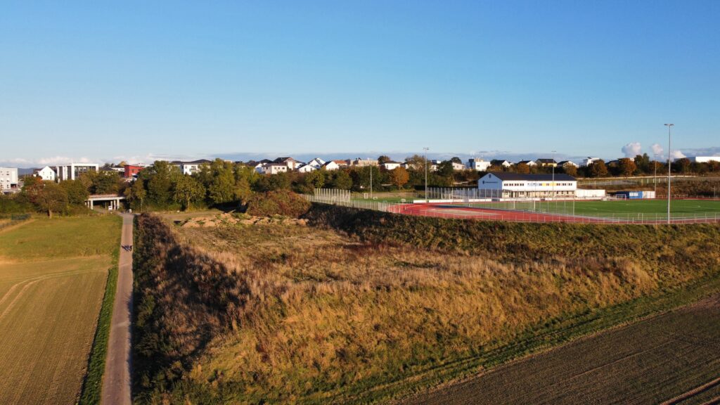 Luftaufnahme des Erdhügels neben dem Sportpark Görsroth: Die großflächige Erdaufschüttung auf Gemeindeland grenzt direkt an die Sportanlage mit Kunstrasenplatz und Tartanbahn. Zwei Spaziergänger auf dem angrenzenden Feldweg verdeutlichen die enorme Dimension der Aufschüttung. Die Fläche liegt seit Jahren ungenutzt brach – eine Baugenehmigung fehlt auch im Februar 2026 noch immer.